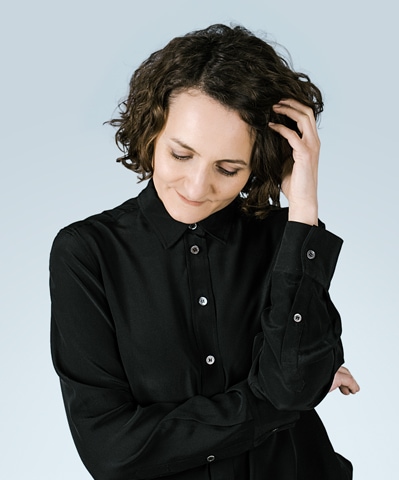 Stefanie Alis Roth in einer schwarzen Bluse posiert mit den Händen auf dem Kopf.